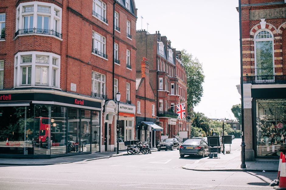 A street scene on Brompton Road in Knightsbridge showing a row of red brick residential buildings with white-framed windows and bay windows on the upper floors. The ground floors house commercial establishments, including a storefront with large glass windows and a black sign reading 'Kartell'. Several parked cars and motorcycles are visible along the curb, and a red double-decker bus is reflected in the shop windows. The street is well-lit with natural daylight, and a Union Jack flag hangs from one of the buildings. The scene conveys a tidy, well-maintained urban area typical of Knightsbridge, with no visible litter or dirt, indicating routine surface cleaning and maintenance by local cleaning services like Carpet Cleaning Knightsbridge.