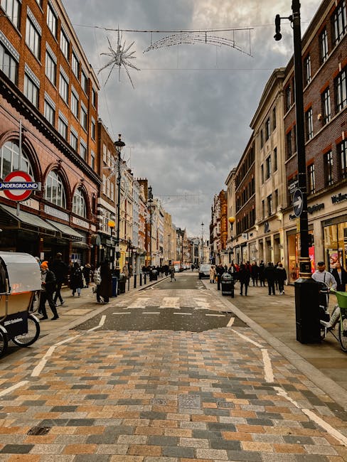 A busy pedestrian street in Knightsbridge with storefronts on both sides, featuring brick and stucco facades with large glass windows and awnings. The cobblestone pavement appears clean and well-maintained, with a section of smoother, lighter-colored stones. Overhead, festive string lights and a decorative star-shaped ornament are suspended across the street. People are walking along the sidewalks and crossing the street, some carrying shopping bags. The scene is bathed in natural daylight with an overcast sky, and the overall atmosphere emphasizes urban cleanliness and vibrant city life, reflecting the professional cleaning standards promoted by Carpet Cleaning Knightsbridge, as seen on their Brompton Road same-day cleaning tips page.