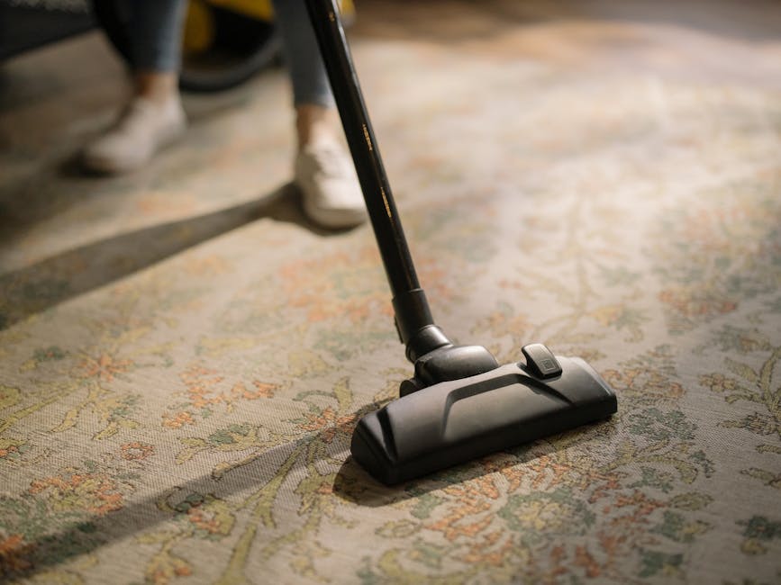 Close-up of a vacuum cleaner head actively cleaning a patterned, plush carpet in a room with soft, natural lighting. The carpet features intricate floral designs in shades of beige, green, and orange. The vacuum's black attachment is pressed directly onto the carpet surface, with a visible switch on top for operation. The background shows part of a person's legs and shoes, suggesting a domestic setting. The image emphasizes surface cleaning and maintenance, showcasing Carpet Cleaning Knightsbridge's professional approach to deep cleaning and hygiene for residential spaces in Knightsbridge.
