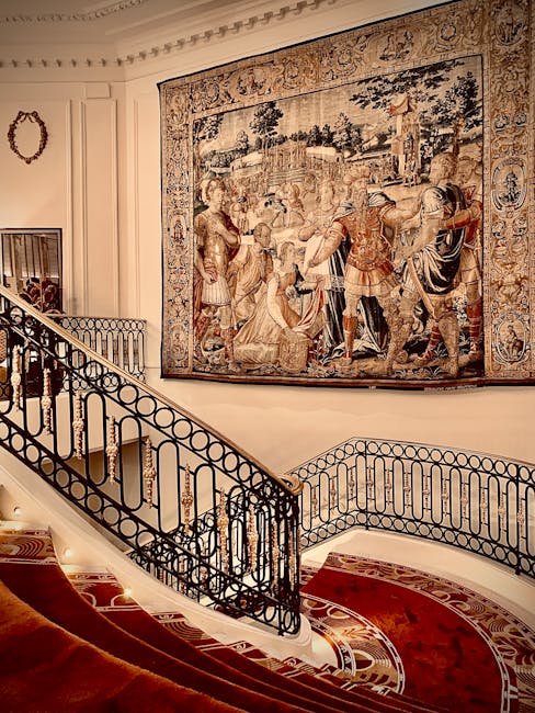 Interior view of a luxurious staircase with a black wrought iron balustrade featuring circular and linear patterns, leading up to a wall adorned with a large, intricate tapestry depicting historical or mythological scenes with figures in classical armor. The staircase is carpeted with a plush red runner edged with a decorative pattern, and the surrounding walls are painted in a soft, neutral tone. On the wall, a small decorative mirror or ornate frame is visible. The lighting is warm and soft, highlighting the fine details of the tapestry and the elegant design of the staircase, creating an atmosphere of classic sophistication. This setting emphasizes the importance of maintaining cleanliness and visual appeal in high-end domestic environments, which is a specialty of Carpet Cleaning Knightsbridge.