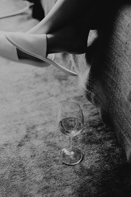 A close-up of a woman's legs resting on a sofa with a visible fabric texture, wearing stylish high heels and stockings. A glass of white wine is placed on a carpeted floor nearby. The setting appears to be a living room with soft lighting and a clean, well-maintained carpet. The image emphasizes a tidy and hygienic domestic space, with Carpet Cleaning Knightsbridge specializing in surface cleaning and sanitisation services for residential environments, as suggested by the page title 'Emergency carpet cleaning for wine stains Knightsbridge' at https://carpetcleaningknightsbridge.co.uk.