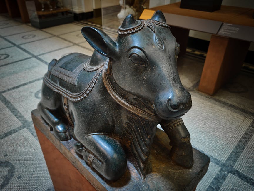 A detailed bronze or similar metal sculpture of a chubby animal, possibly a pig or a boar, displayed on a wooden pedestal in an indoor setting. The sculpture features intricate decorative elements, including a saddle-like adornment on its back and a chain or bead motif around its neck and body. The surface of the sculpture is smooth and polished, with a dark patina that reflects the ambient lighting. The background includes a tiled floor with a geometric pattern and a wooden table or desk, indicating a room dedicated to art, decoration, or a reception area. The lighting is moderate, highlighting the details of the sculpture's craftsmanship, and the area appears clean and well-maintained, in line with professional cleaning standards by Carpet Cleaning Knightsbridge.