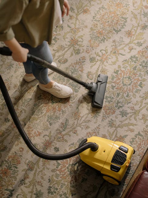 A person is performing surface cleaning on a large, ornate floral-patterned carpet using a vacuum cleaner with a long metal wand, connected to a yellow portable vacuum unit. The scene is set in a well-lit room, highlighting the intricate design and clean condition of the carpet, with the individual wearing beige shoes and casual clothing. The setting appears to be an indoor space, possibly a living room or a commercial area, where professional deep cleaning or maintenance is being carried out, showcasing the services of Carpet Cleaning Knightsbridge during their sanitisation and carpet maintenance process.