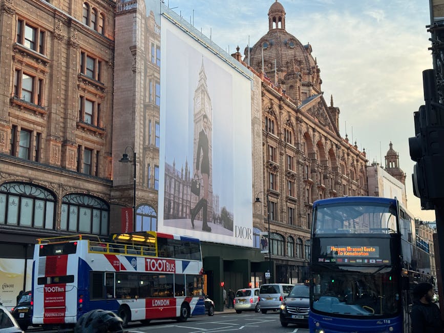 A busy street scene in Knightsbridge, featuring historic ornate buildings with intricate stonework and large windows. In the foreground, there are two double-decker buses, one with a red and white livery labeled 'TOOT Bus' and the other an open-top bus displaying a digital route sign. Pedestrians walk along the sidewalk, and a large outdoor advertisement for Dior hangs on the building, depicting a model in stylish clothing. The scene is illuminated by natural daylight with a clear sky in the background, highlighting the cleanliness and well-maintained appearance of the street and surrounding architecture. The area reflects a blend of historic charm and modern urban activity, with visible signage and traffic indicating a vibrant commercial district. Carpet Cleaning Knightsbridge offers professional surface cleaning and deep cleaning services to maintain this level of hygiene and presentation.