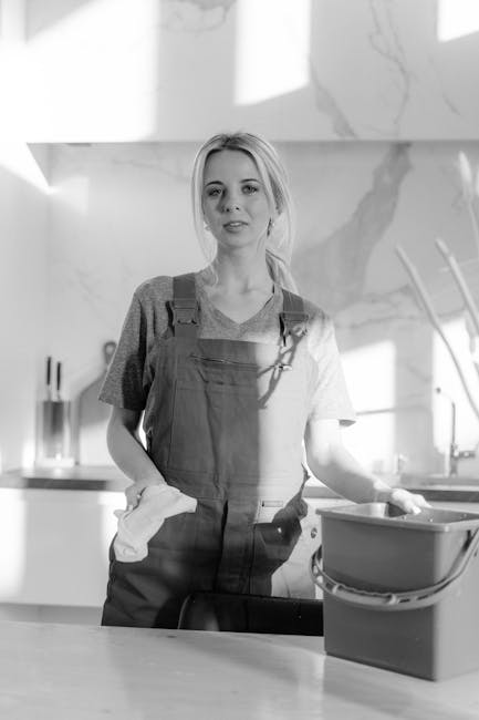 A young woman with light-colored hair tied back, wearing a casual t-shirt and work apron, stands in a modern kitchen holding a cleaning cloth in her right hand and resting her left hand on a stainless steel sink. In front of her is a plastic bucket filled with cleaning supplies. The kitchen features a marble-effect wall, a sleek countertop, and minimalistic utensils in the background, illuminated by natural light from a window. The scene depicts a professional approach to surface cleaning and sanitisation, aligning with deep cleaning and domestic cleaning services promoted by Carpet Cleaning Knightsbridge, emphasizing hygiene and care in residential environments.