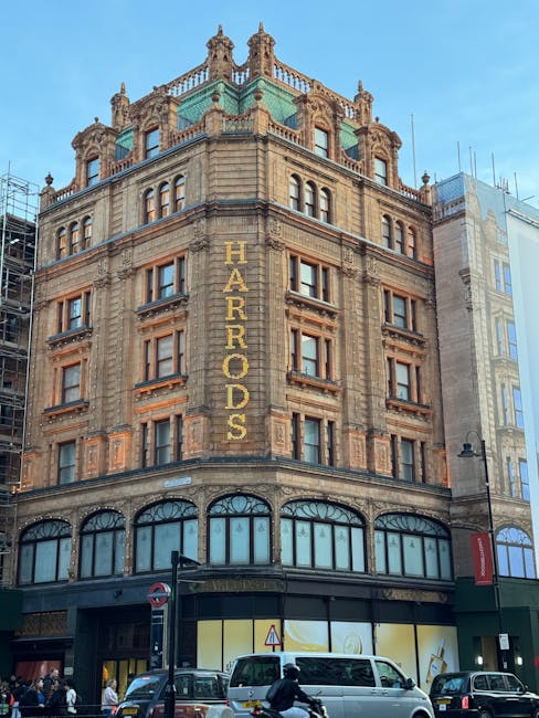 The image depicts a historic multi-story building in Knightsbridge, featuring ornate stonework and large windows on each floor. The building is labeled 'HARRODS' in prominent gold letters along the vertical facade. The ground floor has arched windows and entrances, with a modern glass storefront visible. The structure's upper levels are decorated with decorative cornices, turrets, and spires, demonstrating classic architectural details. The building is set against a clear blue sky, with busy street activity in front, including cars and pedestrians. The scene, captured in daylight, showcases the building’s aged stone exterior that may require regular cleaning and maintenance to preserve its intricate features, aligning with the themes of surface and deep cleaning in line with Kensington and Chelsea council carpet disposal rules. Carpet Cleaning Knightsbridge offers professional cleaning services to help maintain such historic buildings’ hygiene and appearance.
