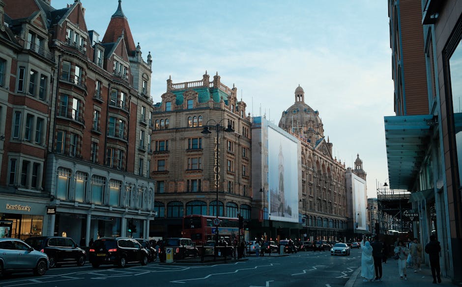 A city street scene in Knightsbridge, London, featuring historic and modern buildings with intricate architectural details, large glass windows, and prominent signage. The street is busy with parked cars along the curb and pedestrians walking on the sidewalk. There are street lamps and a double-decker bus visible in the background, under a clear blue sky with natural daylight illuminating the scene. The surroundings include retail shops, including a visible Guess store, and the overall environment appears clean and well-maintained. Carpet Cleaning Knightsbridge specializes in surface cleaning and deep cleaning services aimed at preserving the hygiene and appearance of residential and commercial spaces in the Knightsbridge area, following local disposal and maintenance regulations.