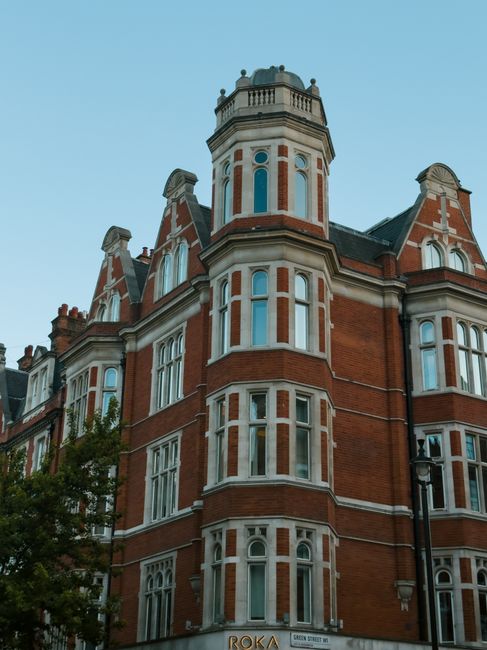 The image depicts a historic multi-story building in Knightsbridge, featuring ornate stonework and large windows on each floor. The building is labeled 'HARRODS' in prominent gold letters along the vertical facade. The ground floor has arched windows and entrances, with a modern glass storefront visible. The structure's upper levels are decorated with decorative cornices, turrets, and spires, demonstrating classic architectural details. The building is set against a clear blue sky, with busy street activity in front, including cars and pedestrians. The scene, captured in daylight, showcases the building’s aged stone exterior that may require regular cleaning and maintenance to preserve its intricate features, aligning with the themes of surface and deep cleaning in line with Kensington and Chelsea council carpet disposal rules. Carpet Cleaning Knightsbridge offers professional cleaning services to help maintain such historic buildings’ hygiene and appearance.