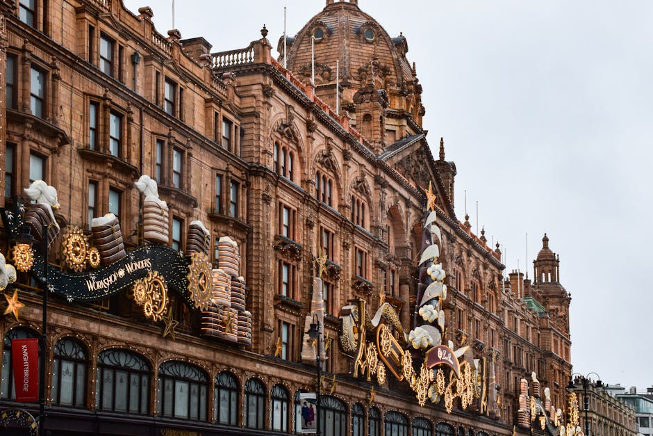Exterior view of a historic sandstone building decorated with festive holiday lights, including illuminated star and gift box ornaments, alongside large caramel-colored panels. The building features arched windows with black frames, ornate stone detailing, and a prominent dome roof with decorative spires, situated in Knightsbridge. The scene is set during daytime under an overcast sky, with the decorations suggesting preparations for a seasonal event or celebration, highlighting a clean, well-maintained facade that reflects the importance of surface cleaning and preservation by Carpet Cleaning Knightsbridge for maintaining the building's appearance during festive times.