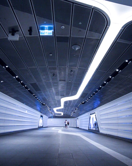 A modern underground corridor with a curved black ceiling featuring integrated LED lighting, creating a bright, clean atmosphere. The walls are lined with white panels with horizontal ridges, contributing to a sleek, hygienic appearance suitable for high-traffic areas. The smooth, gray tiled floor is spotless and reflects the lighting, indicating recent deep cleaning and sanitisation. In the distance, two people are walking, emphasizing the spaciousness of the environment. The setting exemplifies professional surface cleaning and maintenance typical of emergency cleaning services for event stains, as provided by Carpet Cleaning Knightsbridge, situated near Knightsbridge Station.