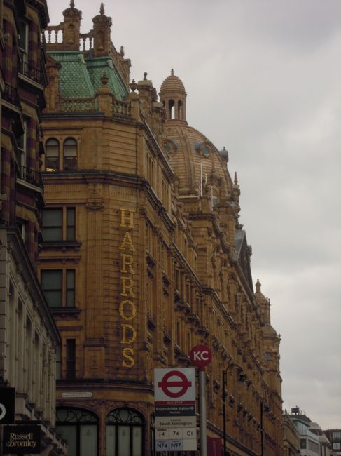 An exterior view of a historic sandstone building in Knightsbridge, featuring ornate architectural details, large windows, and a prominent yellow sign reading 'HARRODS' vertically along the facade. Th