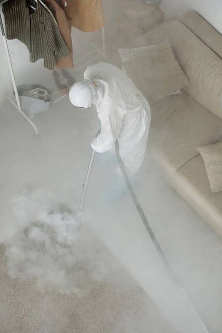 A professional cleaner in protective white clothing and a hood is using a steam or fogging machine for deep cleaning or sanitisation of a cream-colored carpet in a modern living room. The room features a beige upholstered sofa with matching cushions and a light wood floor, visible beneath the mist. Nearby, there is a white drying rack with hanging clothes, and sunlight filters into the space, highlighting the clean and tidy environment. The scene demonstrates surface cleaning and hygiene maintenance in a residential setting, with focus on the equipment used for effective cleaning. Carpet Cleaning Knightsbridge offers expert domestic cleaning services, including upholstery care and stain removal, as seen in this high-quality image.